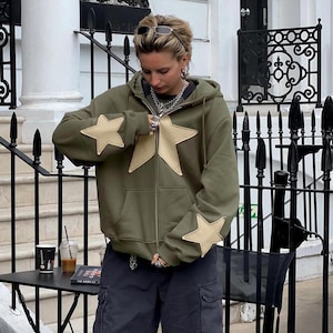 May include: Olive green zip-up hoodie with large tan star appliqués on the front and sleeves. The hoodie has a drawstring hood and a front zipper. The person is wearing dark cargo pants and sunglasses.