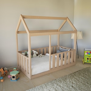 Può includere: Un letto a forma di casa in legno chiaro con un design laterale e del tetto a doghe. Il letto è arredato con una coperta e cuscini bianchi e circondato da peluche. I giocattoli sono sparsi sul pavimento nelle vicinanze.