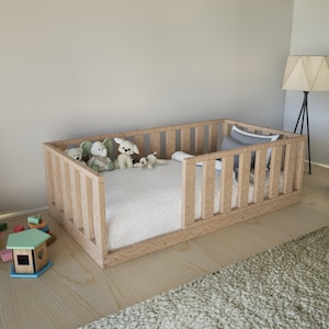 以下が含まれることがあります： 側面が垂直スラットの明るい色の木製幼児用ベッド。ベッドには白いマットレスといくつかのぬいぐるみが入っています。小さな木製のおもちゃの家とブロックが床にあります。