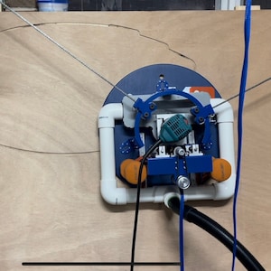 May include: A woodworking machine with a blue and white frame, orange components, and a green power tool is mounted on a wooden surface. A black hose and a blue cable extend from the machine. A thin wire follows a curved line on the wood.