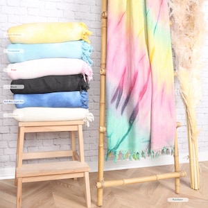 May include: A stack of colorful, fringed blankets in light yellow, turquoise, pink, black, dark blue, and natural. A rainbow-colored blanket hangs on a bamboo ladder. The blankets are displayed on a wooden step stool and ladder.
