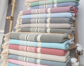 May include: A stack of colorful Turkish towels with fringe edges. The towels are folded and stacked in a pyramid shape. The towels are made of cotton and have a variety of patterns, including stripes, herringbone, and chevron.