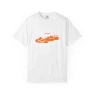 Op de afbeelding: Wit T-shirt met een oranje illustratie van een raceauto en het McLaren-logo. Het shirt heeft een ronde hals en korte mouwen. Het ontwerp is gecentreerd op de voorkant. De auto is in Formule 1-stijl.