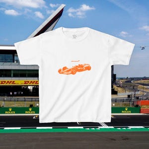 May include: White t-shirt featuring an orange illustration of a race car with the word "McLaren" above it. The shirt is set against a backdrop of a race track and blue sky.