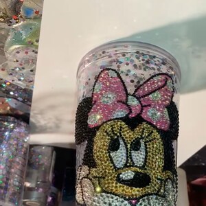 May include: A clear plastic tumbler with a Minnie Mouse design. The design is made of rhinestones in black, pink, gold, and white. The tumbler is filled with glitter.