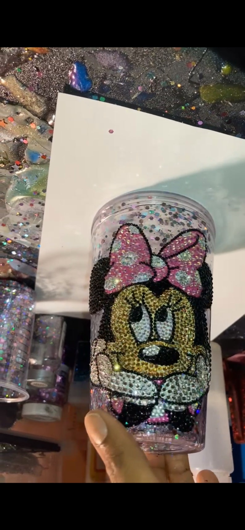 May include: A clear plastic tumbler with a Minnie Mouse design. The design is made of rhinestones in black, pink, gold, and white. The tumbler is filled with glitter.