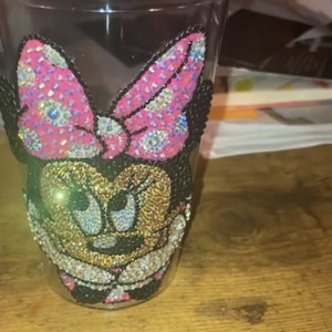 May include: A clear plastic tumbler with a rhinestone design of Minnie Mouse. The design features a pink bow, black ears, and a gold face.