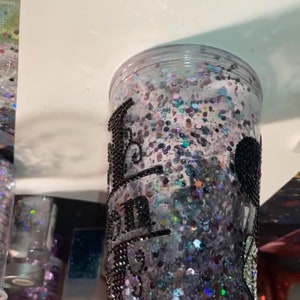 May include: A clear plastic tumbler with a black glitter design and the words "Bad to the Bone" written in black. The tumbler is filled with iridescent glitter.