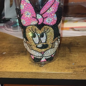 May include: A clear plastic tumbler with a rhinestone design of Minnie Mouse. The design features a pink bow with white polka dots and black ears.
