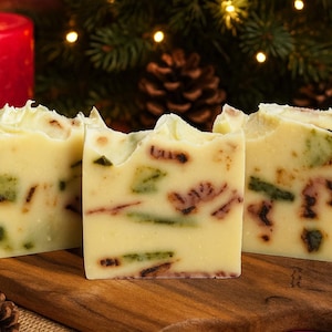 May include: Three bars of handmade soap with a pale yellow base, containing green and brown pieces. The soaps are on a wooden board, with a lit red candle and pine cones in the background, suggesting a festive theme.
