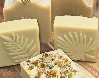 May include: Five handmade soaps in varying shapes and sizes. Some bars are topped with dried flowers. Two bars feature leaf imprints. The soaps are light yellow and beige, and rest on a wooden surface.