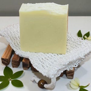 May include: A rectangular bar of light green soap with a textured white top, resting on a wooden soap dish with a white mesh soap saver. The soap dish is made of dark brown wood. Green leaves and white flowers are scattered around the soap.