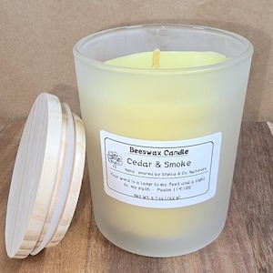 May include: A frosted glass Beeswax Candle with a wooden lid. The candle is labeled "Cedar & Smoke" and has a net weight of 5.7 oz (162 g). The candle is hand-poured by Stella & Co. Naturals.