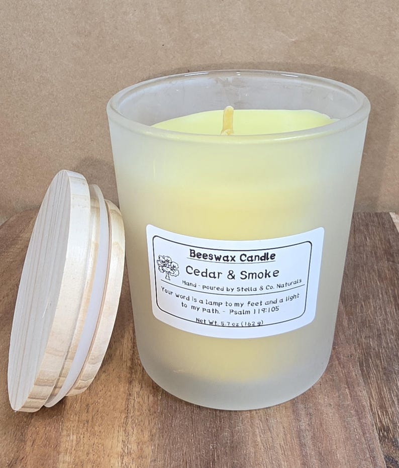 May include: A frosted glass Beeswax Candle with a wooden lid. The candle is labeled "Cedar & Smoke" and has a net weight of 5.7 oz (162 g). The candle is hand-poured by Stella & Co. Naturals.