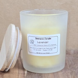 May include: A frosted glass jar candle with a wooden lid, labeled "Beeswax Candle Lavender." The candle is yellow and has a label with text including "Net Wt. 5.1 oz (146 g)." The lid is partially open, resting on a wooden surface.