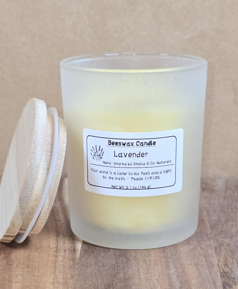 May include: A frosted glass jar candle with a wooden lid, labeled "Beeswax Candle Lavender." The candle is yellow and has a label with text including "Net Wt. 5.1 oz (146 g)." The lid is partially open, resting on a wooden surface.