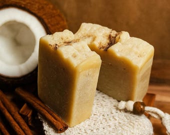 May include: Two rectangular bars of handmade soap with a light beige color and a textured surface, resting on a white loofah. Cinnamon sticks, vanilla beans, and a coconut half are arranged around the soap on a wooden surface.