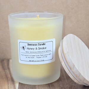 May include: A frosted glass beeswax candle labeled "Honey & Smoke" with a wooden lid. The candle has a yellow hue and a label with text including "Beeswax Candle" and "Net Wt. 6.1 oz (174 g)." The lid is partially open.