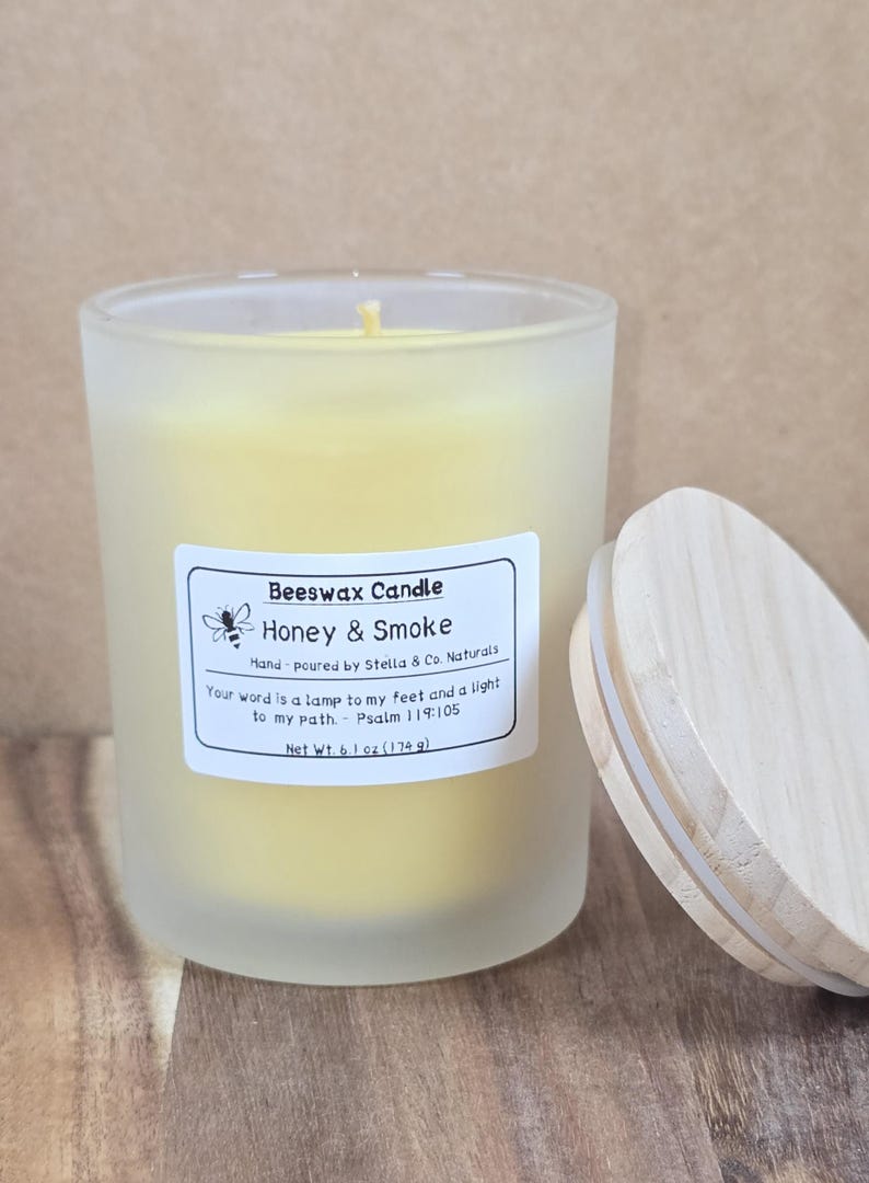 May include: A frosted glass beeswax candle labeled "Honey & Smoke" with a wooden lid. The candle has a yellow hue and a label with text including "Beeswax Candle" and "Net Wt. 6.1 oz (174 g)." The lid is partially open.