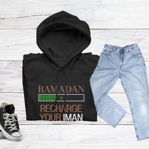 Puede incluir: Sudadera con capucha negra con un gr&aacute;fico verde y marr&oacute;n que dice "Ramadan Recharge Your Iman". La sudadera se combina con jeans azules y zapatillas negras.