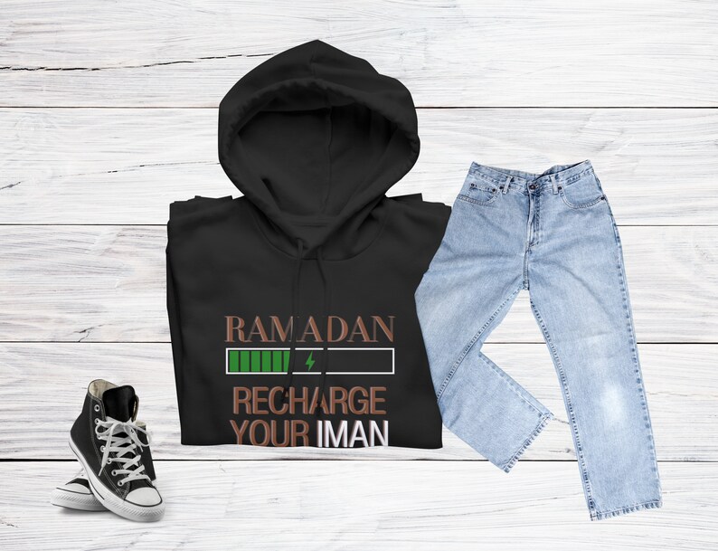 Puede incluir: Sudadera con capucha negra con un gr&aacute;fico verde y marr&oacute;n que dice "Ramadan Recharge Your Iman". La sudadera se combina con jeans azules y zapatillas negras.