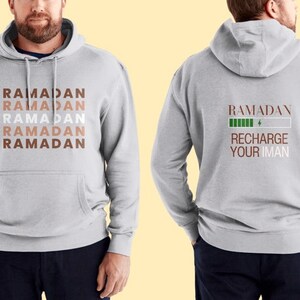 Puede incluir: Una sudadera gris con la palabra "RAMADAN" repetida cinco veces en diferentes tonos de marr&oacute;n en la parte delantera. La parte posterior de la sudadera tiene la palabra "RAMADAN" con una barra de bater&iacute;a verde y el texto "RECHARGE YOUR IMAN".
