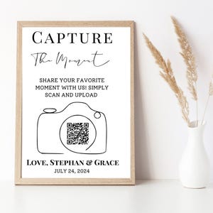 May include: A black and white printable sign with the text "CAPTURE The Moment" and a stylized camera with a QR code. The text below the camera reads "SHARE YOUR FAVORITE MOMENT WITH US! SIMPLY SCAN AND UPLOAD LOVE, STEPHAN & GRACE JULY 24, 2024".
