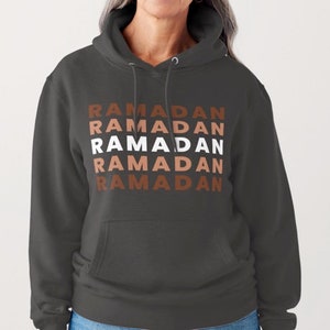 Puede incluir: Una sudadera con capucha gris oscuro con la palabra "RAMADAN" repetida cinco veces en un patr&oacute;n apilado. El texto est&aacute; en blanco con acentos marrones y naranjas.