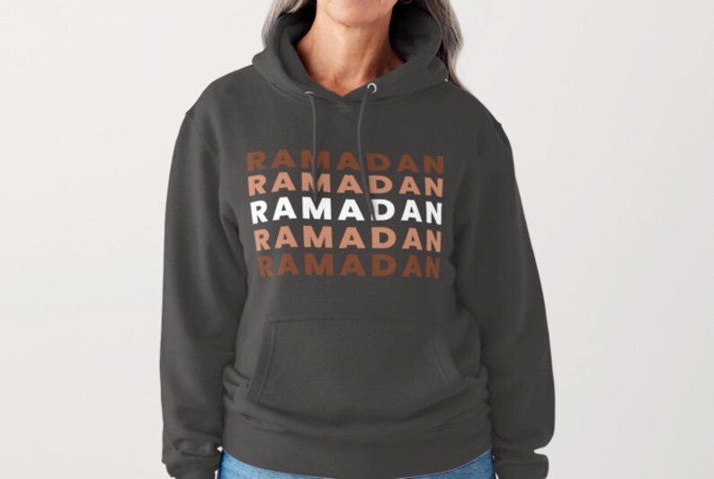 Puede incluir: Una sudadera con capucha gris oscuro con la palabra "RAMADAN" repetida cinco veces en un patr&oacute;n apilado. El texto est&aacute; en blanco con acentos marrones y naranjas.