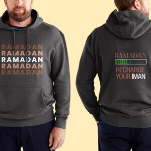 Puede incluir: Una sudadera con capucha gris oscuro con un bolsillo en la parte delantera. La parte delantera de la sudadera tiene la palabra "RAMADAN" repetida cinco veces en un patr&oacute;n apilado. La parte posterior de la sudadera tiene las palabras "RAMADAN RECHARGE YOUR IMAN" con una barra de progreso verde debajo de la palabra "RAMADAN".