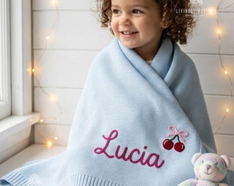 May include: A light blue knitted blanket featuring the name Lucia embroidered in dark pink script lettering. The blanket includes a small embroidered cherry motif with a pink bow and is displayed alongside a pastel-colored plush teddy bear.