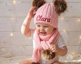 May include: A light pink ribbed knit beanie with two large brown faux fur pom-poms and the name Clara embroidered in red script. The set includes a matching light pink ribbed knit scarf featuring two brown faux fur pom-poms at the ends.