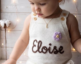 May include: An off-white knitted romper featuring the name Eloise embroidered in dark brown yarn across the chest. The garment includes two light brown wooden buttons on each shoulder strap and a small, textured light purple flower applique.