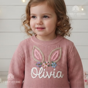 May include: A pink knitted sweater with embroidered bunny ears and a floral design. The name "Olivia" is embroidered in white script below the ears. The sweater has a ribbed texture and is designed for a child.