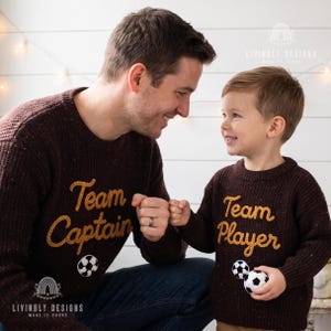 Peut inclure: Deux pulls en tricot marron. L'un porte l'inscription "Team Captain" avec un ballon de foot, l'autre "Team Player" avec deux ballons. Les pulls sont de Livingly Designs. L'image montre un père et son enfant.