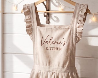 May include: A beige apron hanging on a wooden hanger, featuring ruffled shoulder straps and a gathered skirt. The front is embroidered with the text Valerie's KITCHEN in a dark brown script and block font, with two visible pockets at the bottom.