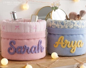 May include: Two round fabric storage baskets with quilted textures. The pink basket features the name Sarah in purple yarn, while the blue basket has a floral rim and the name Arya in yellow yarn. Both baskets are used for organizing small items.