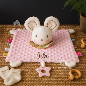 May include: A pink baby security blanket with a plush bunny head and ears. The blanket has a textured surface and colorful ribbons around the edges. The name "Isla" is embroidered on the blanket. Includes a star-shaped teether and baby socks.