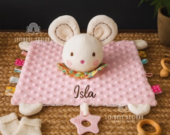 May include: A soft pink baby security blanket featuring a plush mouse head with large ears, a colorful floral collar, and the name Isla embroidered in dark brown. The blanket has a textured dot pattern and a light pink star-shaped plastic teether.