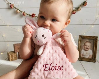 May include: A soft, light pink plush security blanket featuring a stuffed rabbit head with embroidered closed eyes and a small nose. The fabric has a textured dimpled pattern and is personalized with the name Eloise embroidered in dark red.