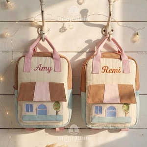 Puede incluir: Dos mochilas beige y marrón con correas rosas y los nombres "Amy" y "Remi" bordados. Cada mochila presenta un diseño de casa con ventanas azules, un tejado marrón y un árbol.