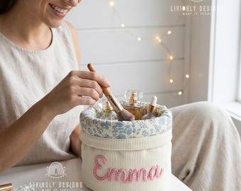 May include: A round, cream-colored corduroy storage basket featuring the name Emma embroidered in pink yarn on the front. The interior is lined with a blue and white floral fabric, and the basket contains several glass perfume bottles.