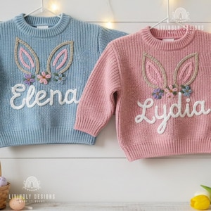 May include: Two children's sweaters, one pale blue and one pink, each with a bunny ear design and a name embroidered on the front. The bunny ears are outlined in a beige colour and decorated with small flowers. The pale blue sweater says "Elena" and the pink sweater says "Lydia".