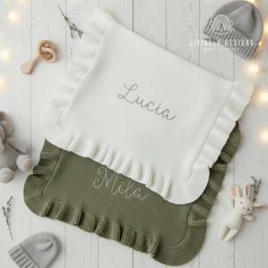Puede incluir: Dos mantas para bebé, una blanca y otra verde oliva, con bordes con volantes. La manta blanca está bordada con el nombre "Lucia", y la verde tiene "Mila". Otros artículos para bebés incluyen un gorro gris, una sonaja de madera y un conejito de peluche.