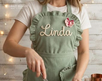 May include: A sage green corduroy apron featuring a ruffled trim along the edges, a white embroidered name "Linda" in cursive script, and a small pink and white butterfly appliqué. The apron is worn over a plain white short-sleeved t-shirt.