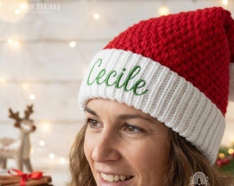 May include: A red and white knit hat featuring a textured red crown and a wide white ribbed cuff. The name Cecile is embroidered in green cursive script on the front of the white cuff. The hat has a classic holiday color scheme.