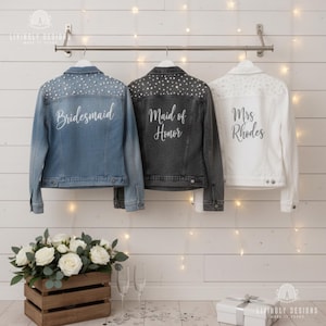 Op de afbeelding: Drie spijkerjacks hangen aan een metalen stang. De jacks zijn lichtblauw, zwart en wit. Op het lichtblauwe jack staat "Bridesmaid", op het zwarte jack "Maid of Honor" en op het witte jack "Mrs. Rhodes". Elk jack heeft decoratieve versieringen.