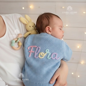 Puede incluir: Un bebé con un suéter de punto azul claro con el nombre "Flora" bordado en colores pastel. También se ven un conejito de crochet amarillo y un mordedor de madera.