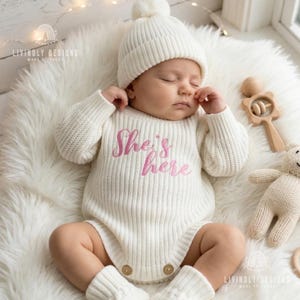 Puede incluir: Un bebé recién nacido con un body de punto blanco con el texto bordado "She's here", un gorro y calcetines a juego. El bebé está acostado sobre una superficie blanca y esponjosa, con un sonajero de madera y un pequeño oso de peluche cerca.