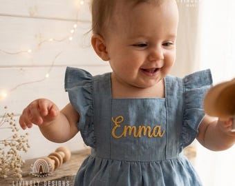 May include: A light blue linen romper featuring ruffled sleeves and the name Emma embroidered in gold cursive thread on the chest. The garment has a square neckline and a gathered waistline, providing a simple and functional design for infants.